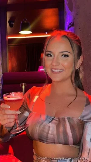 Woman in striped crop top holding drink, smiling in dimly lit indoor setting with red lighting.