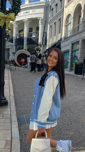 Woman in blue and white varsity jacket and white shorts, holding white handbag, standing on