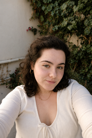 Woman with curly dark hair in white top, outdoors near greenery.