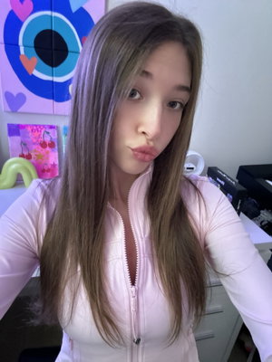 Woman with long brown hair in pink zip-up top taking selfie indoors.