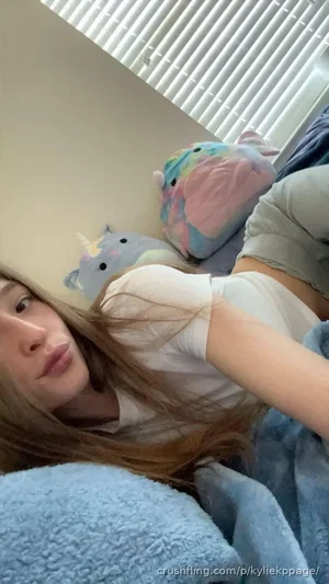 Woman with long hair lying in bed, wearing white top and gray shorts, surrounded by plush toys.