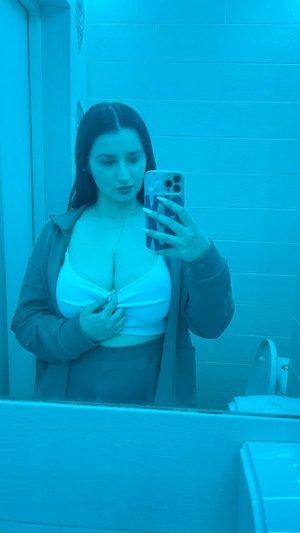 Woman in white top and jacket taking mirror selfie in bathroom.