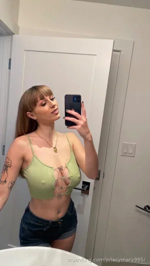 Woman with tattoos wears green crop top and denim shorts, taking mirror selfie in bathroom.