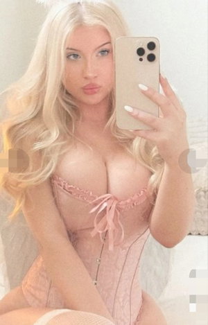 Blonde woman in pink corset taking mirror selfie in bedroom.