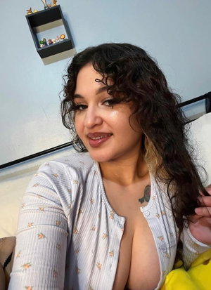 Curvy woman with curly dark hair in floral top taking bedroom selfie.