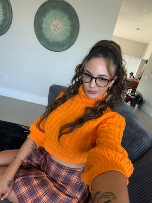 Woman in orange sweater and plaid skirt taking indoor selfie.