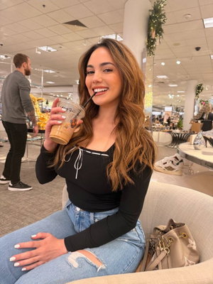 Woman with long brown hair in black top and jeans holding coffee indoors.
