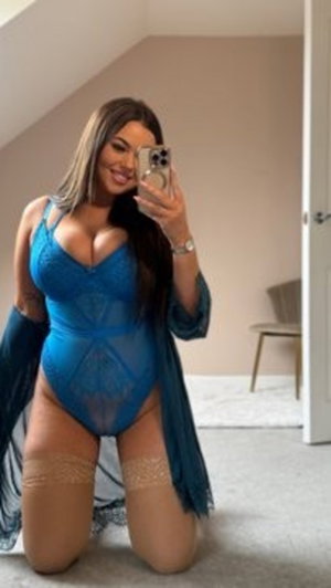 Woman in blue lace bodysuit and sheer robe kneeling indoors, holding phone for mirror selfie.
