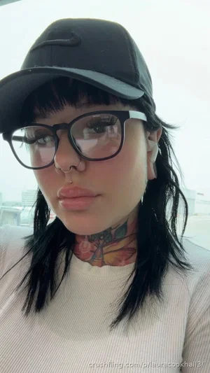 Woman with black hair, glasses, cap, and neck tattoo wearing a white top indoors.
