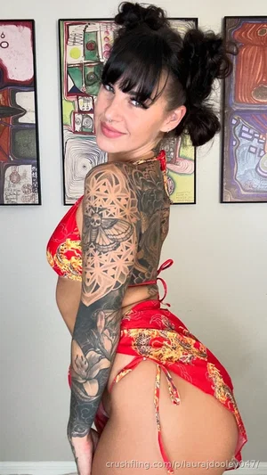 Woman with dark hair in buns, tattoos on arm, wearing red patterned lingerie, posing indoors.