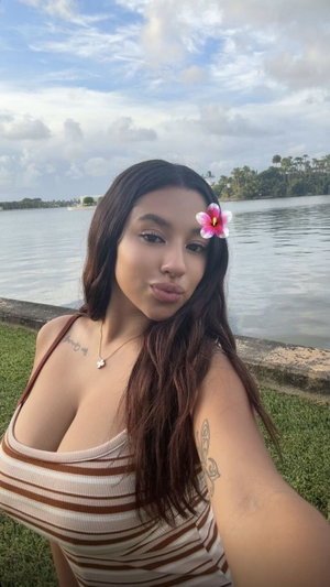Woman with long dark hair and flower in hair, wearing striped top, posing by water.