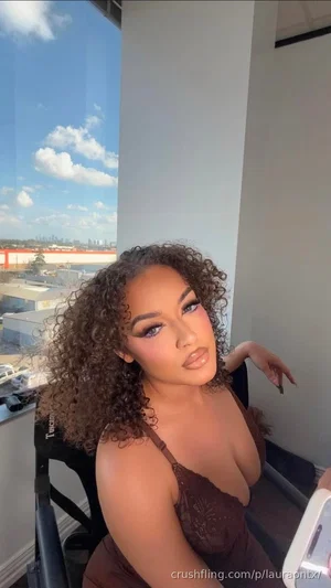 Woman with curly hair in a brown lace top, sitting indoors near a window with city view.