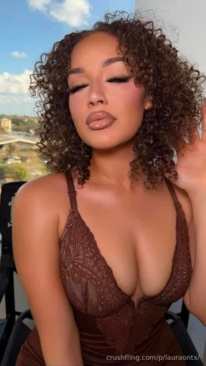 Woman with curly hair in brown lace lingerie, posing outdoors with city view.