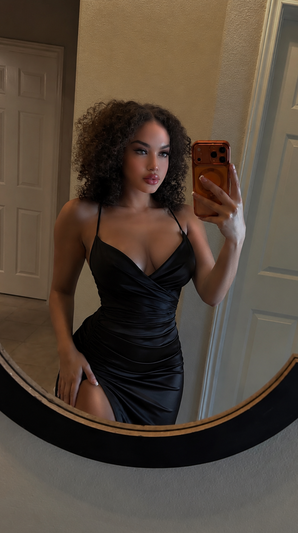 Curly-haired woman in black dress taking mirror selfie indoors.