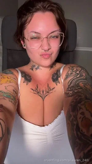 Woman with tattoos, glasses, and a white top, taking a selfie indoors.