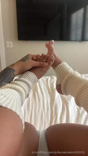 Woman in white socks and underwear on bed, holding foot, tattooed arm visible.