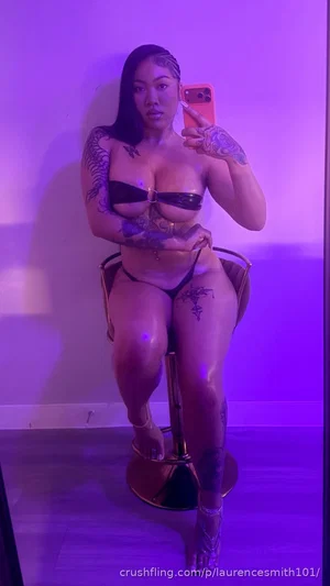 Woman with tattoos in black lingerie sitting on a stool under purple lighting.