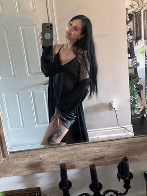 Woman with long dark hair and glasses takes mirror selfie in black sheer robe, showing tattoos on