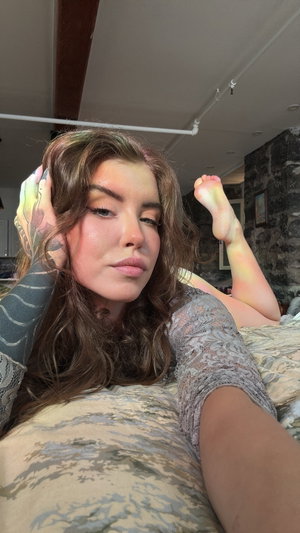 Woman with long brown hair lying on bed, wearing patterned top, tattoo on arm.