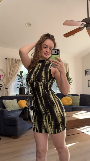 Woman in black and green patterned dress taking mirror selfie in living room.
