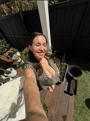 Woman in checkered top taking selfie outdoors on wooden deck with plants.