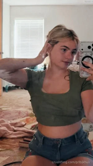 Woman in green crop top and denim shorts taking mirror selfie in bedroom.