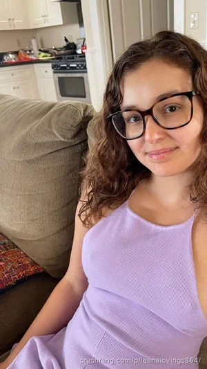 Woman with curly hair and glasses in a purple tank top sitting on a couch in a kitchen.