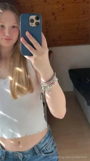 Woman with long blonde hair in white crop top and jeans, taking mirror selfie indoors.