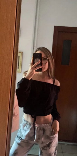Woman in off-shoulder top and tie-dye pants takes mirror selfie indoors.