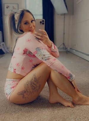 Woman in pink patterned pajamas with tattoo on thigh, sitting on floor taking mirror selfie.