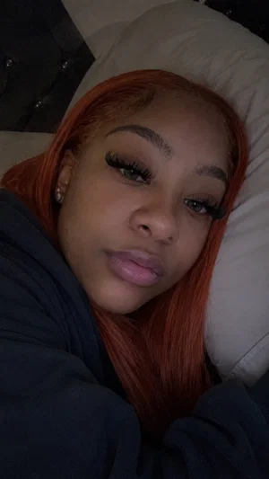Woman with red hair and makeup lying on a bed, wearing a dark top.