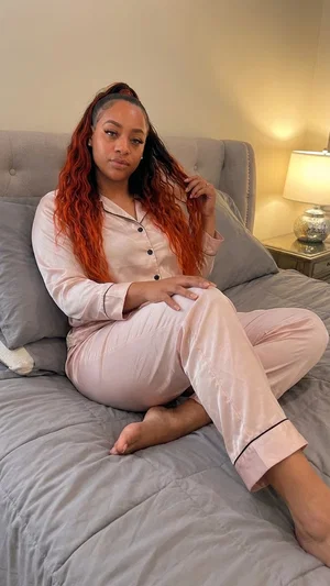 Woman with red hair in pink pajamas sitting on a bed indoors.