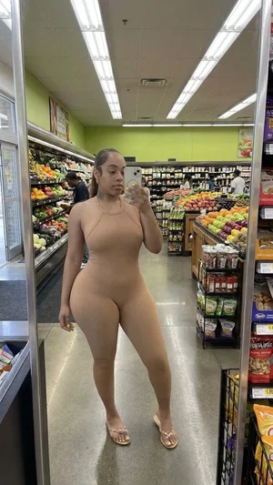 Woman in beige bodysuit taking mirror selfie in grocery store produce aisle.