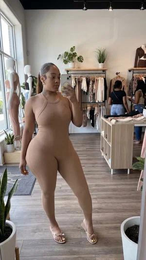 Woman in beige bodysuit and heels taking mirror selfie in clothing store.