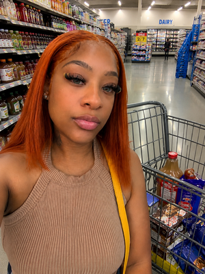 Woman with orange hair in a beige tank top taking a selfie in a grocery store aisle.