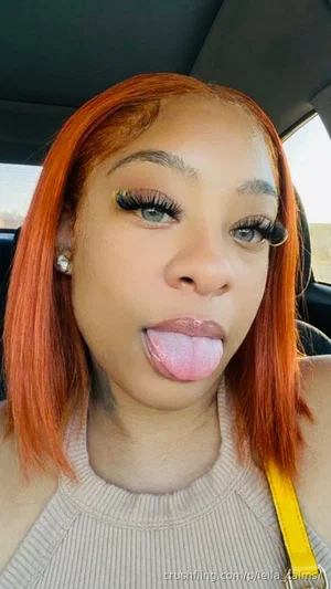 Woman with orange hair and makeup, wearing a beige top, sticking tongue out in car.