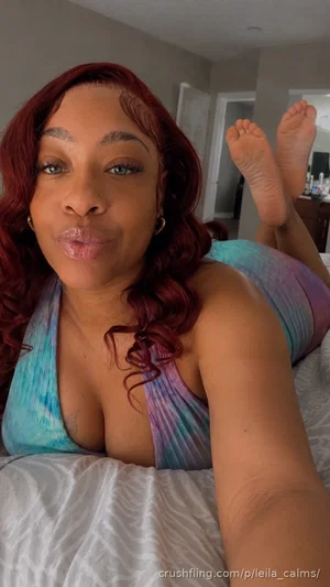 Woman with red hair in colorful top lying on bed, legs raised, indoor setting.