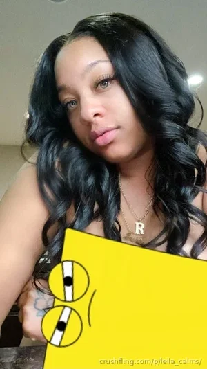 Woman with long black wavy hair, wearing a necklace with an 'R' pendant, holding a yellow object