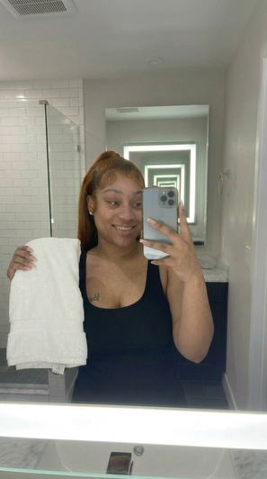 Woman in black tank top holding towel, taking mirror selfie in bathroom.