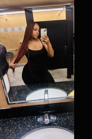 Woman with long braids taking mirror selfie in bathroom wearing black dress.