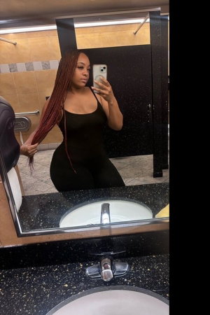 Woman with long braids in black outfit taking mirror selfie in bathroom.