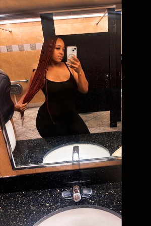 Woman with long braids in black dress taking mirror selfie in bathroom.