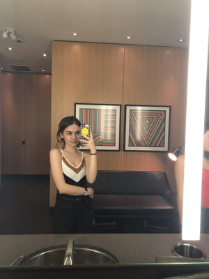 Woman in black and white top and black pants takes mirror selfie in modern room with art on wall.