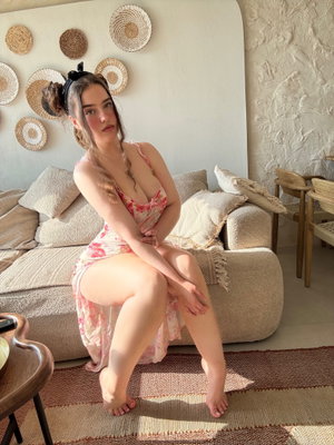 Woman in floral dress sits on couch, legs crossed, in cozy living room with woven wall decor.