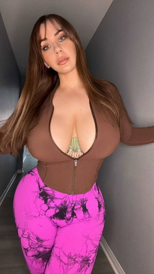 Curvy woman with long brown hair in brown top and pink leggings indoors.