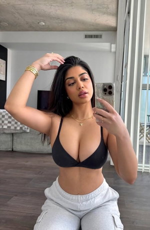 Woman in black bra and gray sweatpants taking mirror selfie in living room.
