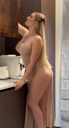 Woman with long blonde hair in beige dress standing in kitchen, side profile.