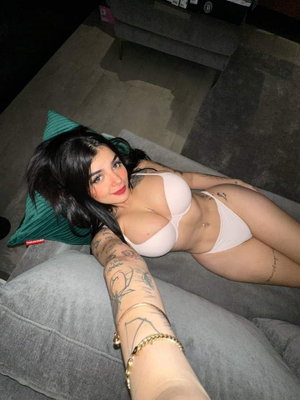 Woman with tattoos in white lingerie lying on gray couch, holding phone for selfie.