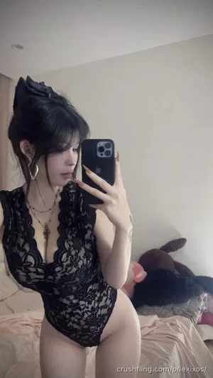 Woman in black lace bodysuit takes mirror selfie in bedroom.