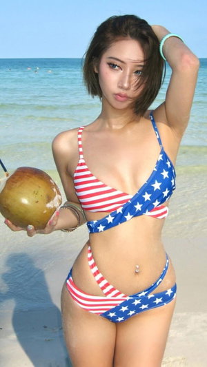 Woman in American flag bikini holding coconut on beach.